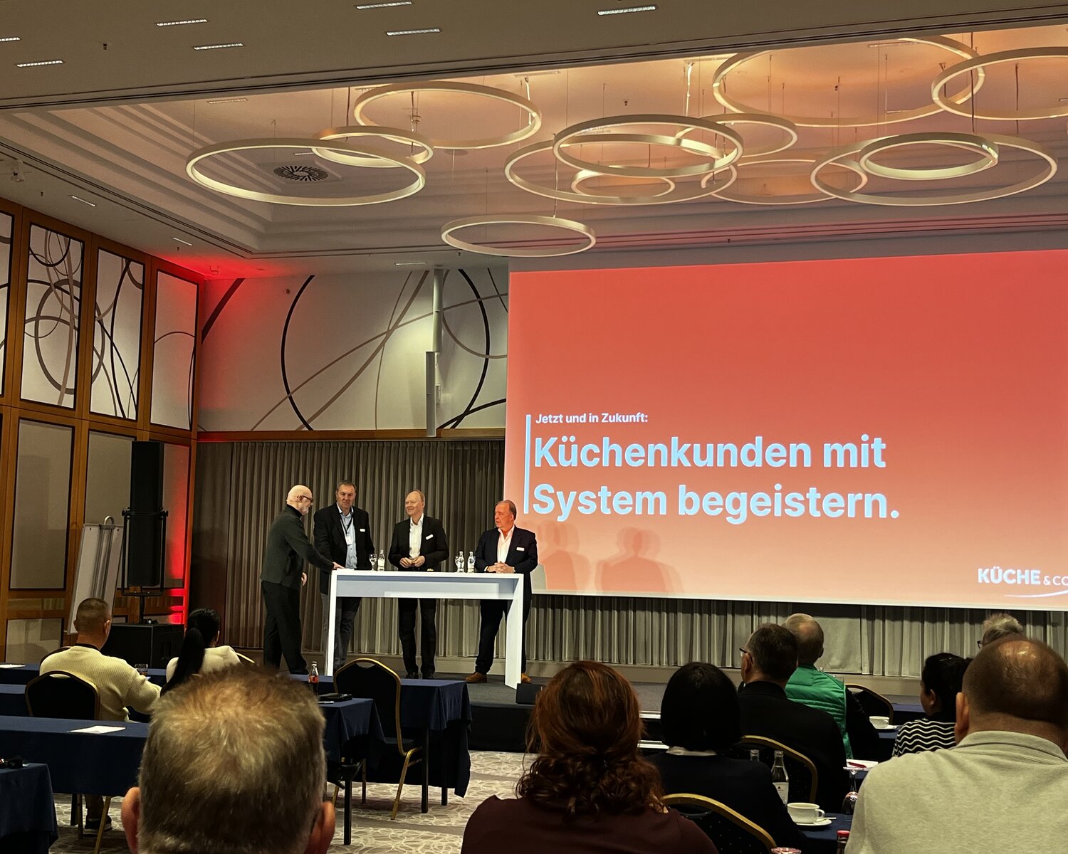 Talkrunde auf der Bühne der Küche&Co-Franchisetage 2025 vor der Leinwand mit dem Claim „Küchenkunden mit System begeistern."